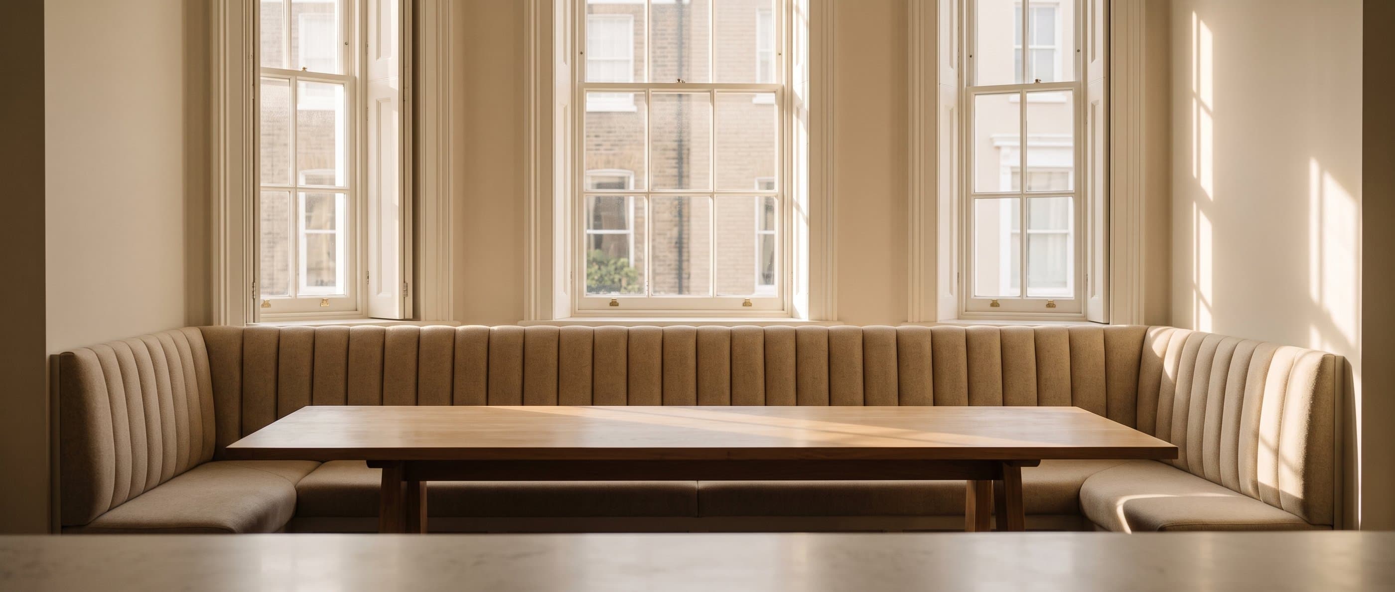 A U-shaped fluted banquette beneath three sash windows, around a long oak table